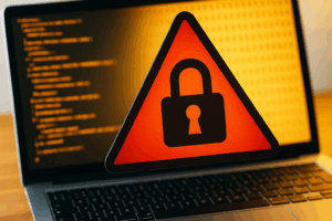 Photo-realistic image of a red cybersecurity warning sign featuring a padlock icon, displayed in front of a laptop screen with coding and binary data, symbolising digital threats to UK businesses.